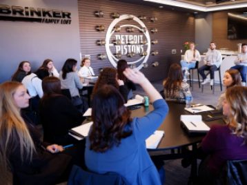 UM students sit at round tables, with Detroit Pistons employees sitting at the far end of the room. One student raises her hand.